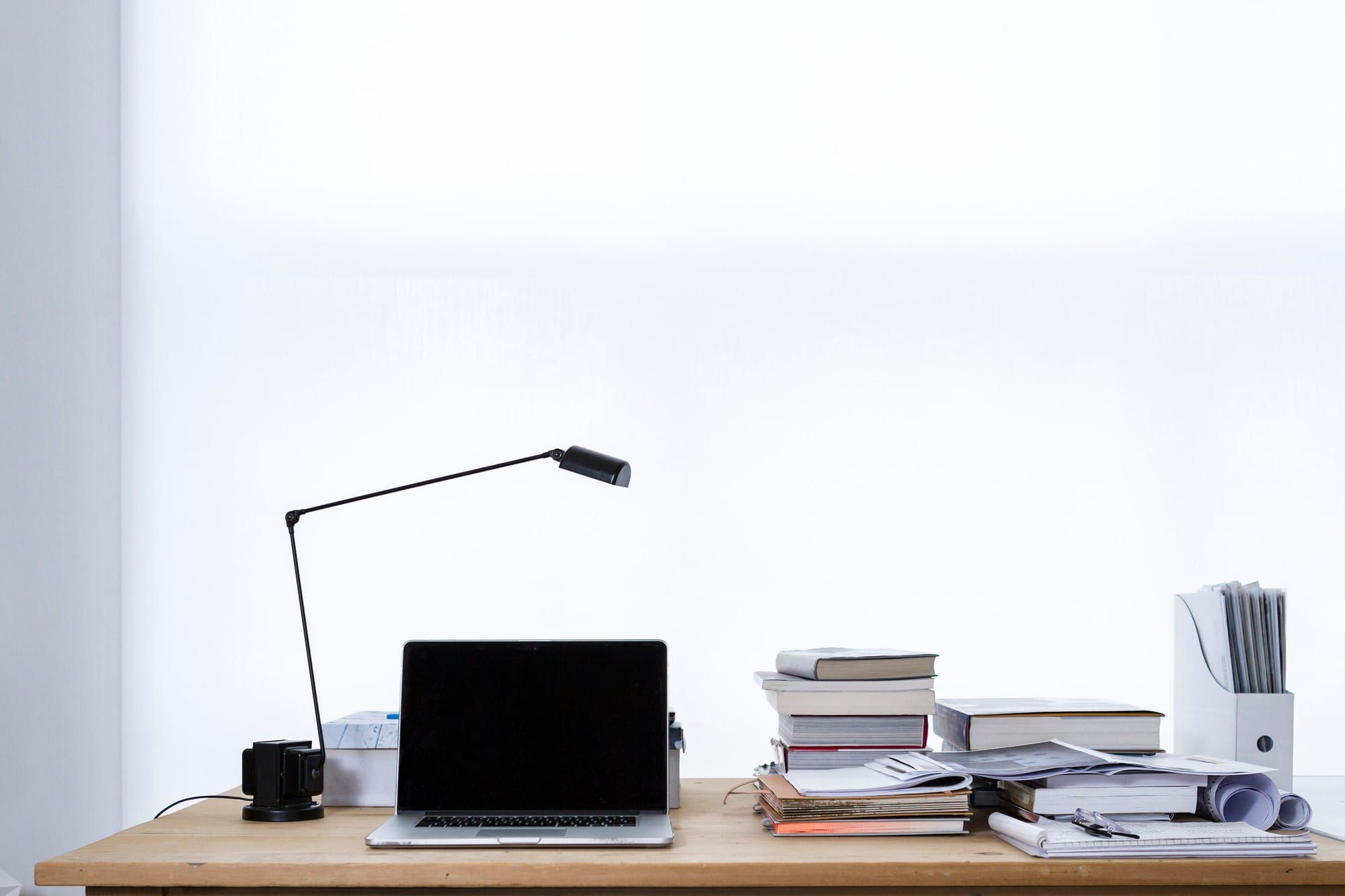 A laptop on a wooden desk beside a black desk lamp and stacks of books against a white wall