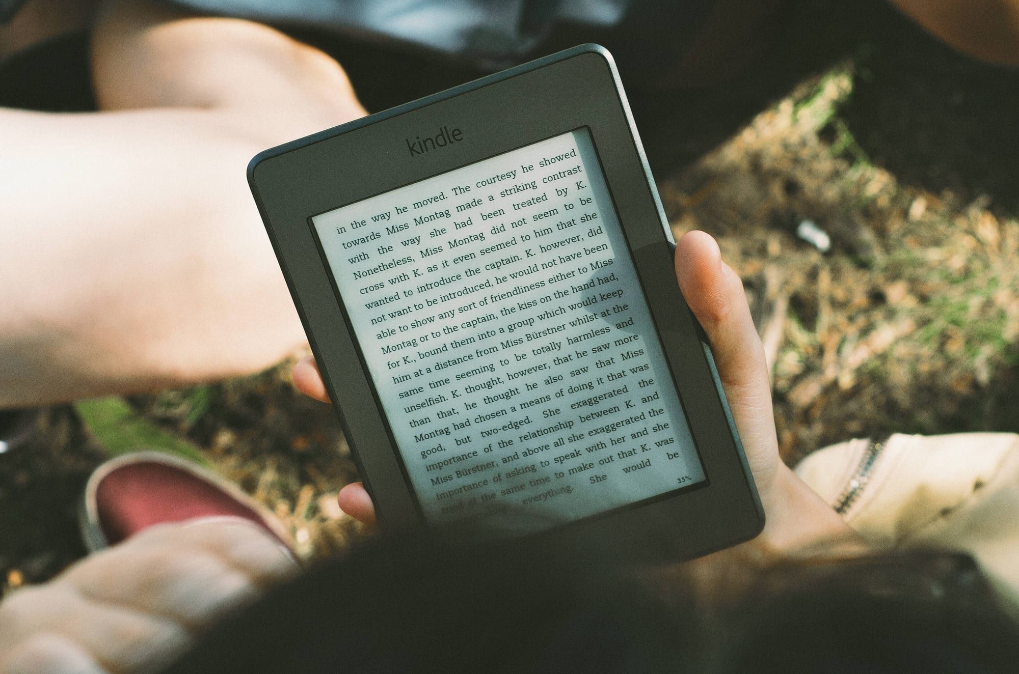 A hand holding a Kindle e-reader showing a page of text, with grass and sandals visible in the background outdoors