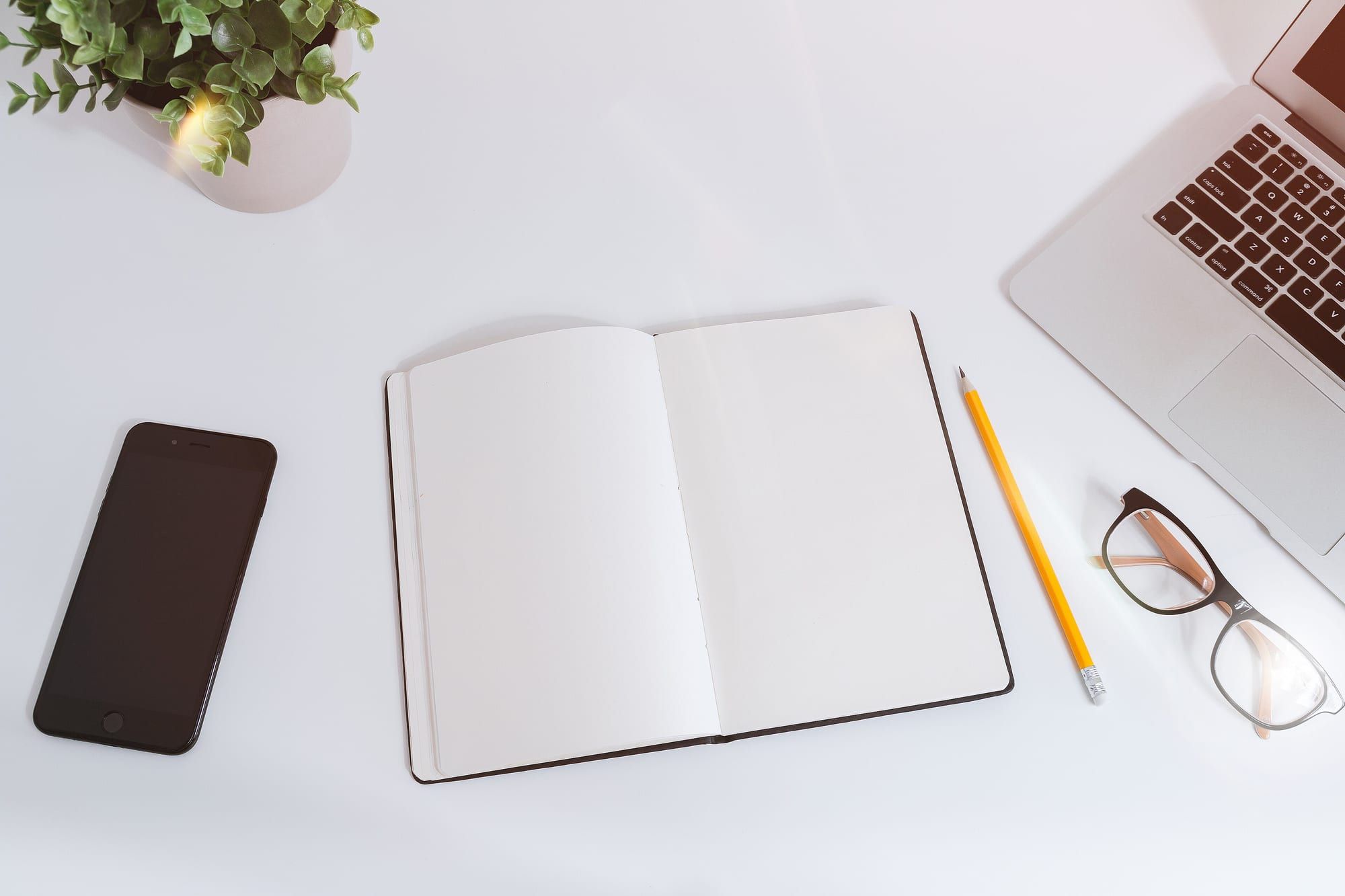 An open blank notebook with a yellow pencil, glasses, a smartphone and a plant on a white desk