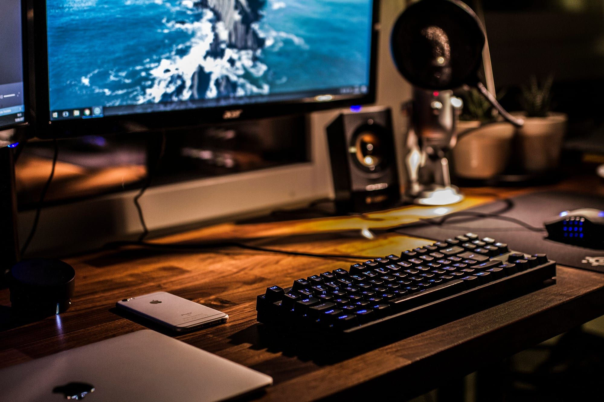 A desktop workstation with a monitor, speakers and a backlit keyboard on a wooden desk in warm light