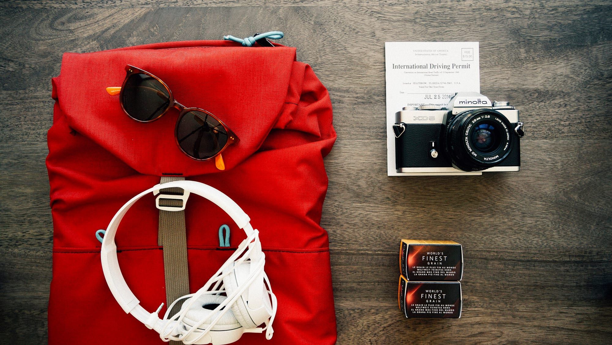 A red backpack on a wooden table beside sunglasses, headphones and a film camera