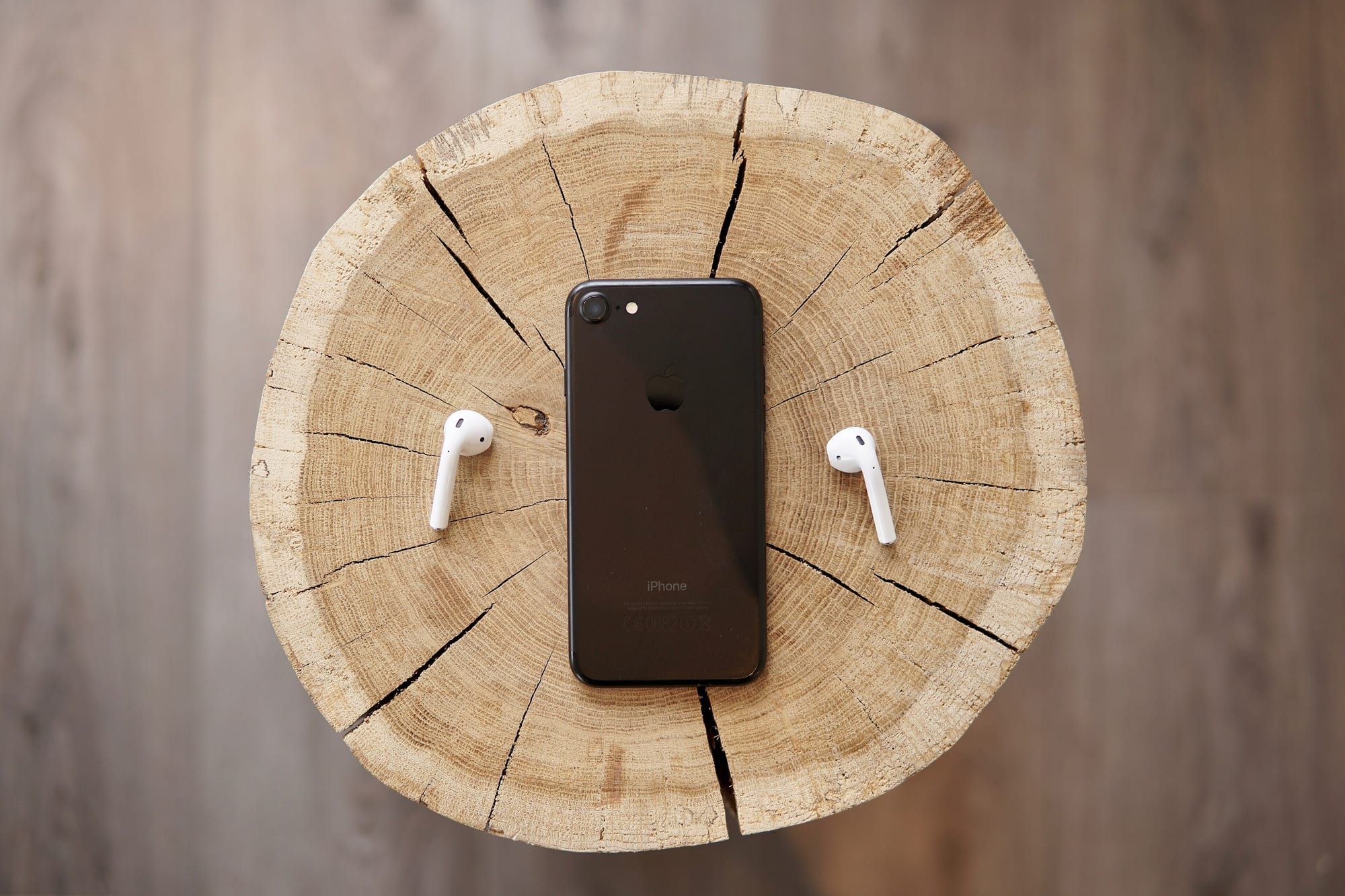 A pair of white wireless earbuds placed either side of a black iPhone on a round wooden slice
